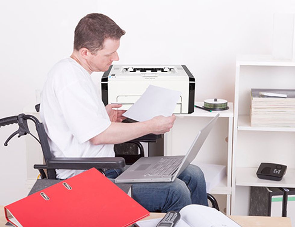 "Foto de um homem em uma cadeira de rodas segurando um pedaço de papel com seu laptop perto de uma impressora."